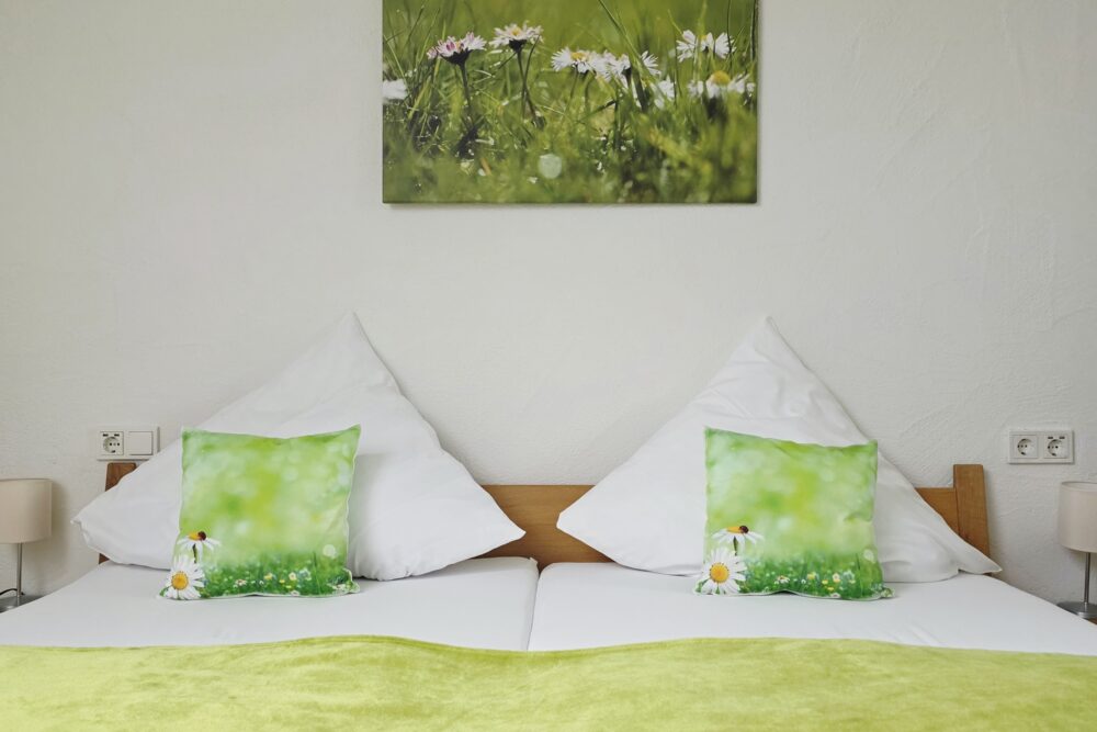 Blick im Hotelzimmer Gänseblümchen auf das Bett mit darüber hängendem Fotodruck von Gänseblümchen auf der Wiese