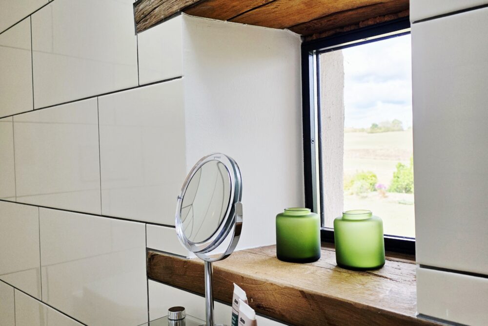 Blick auf das kleine Fenster mit Fernblick im Badezimmer des Hotelzimmers Gänseblümchen, Glasregal mit Produkten und Handtüchern im Vordergrund